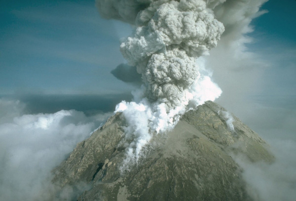 A vigorous eruption column rising over the summit of 1,282-m (4,206 ft)-high Augustine Volcano. Photograph by M.E. Yount, U.S. Geological Survey, March 31, 1986. The image shows a dramatic aerial view of Mount Spurr, an active volcano, during an eruption. A massive plume of light gray ash and smoke billows upward from the volcano's peak in a dense column. The volcano itself appears as a rocky, triangular mountain formation rising above a layer of clouds. The mountain has a grayish-brown color with some snow or ash visible on its slopes. The eruption cloud stands out starkly against a clear blue sky. This powerful display of volcanic activity shows the tremendous force of the erupting volcano as it releases ash high into the atmosphere.