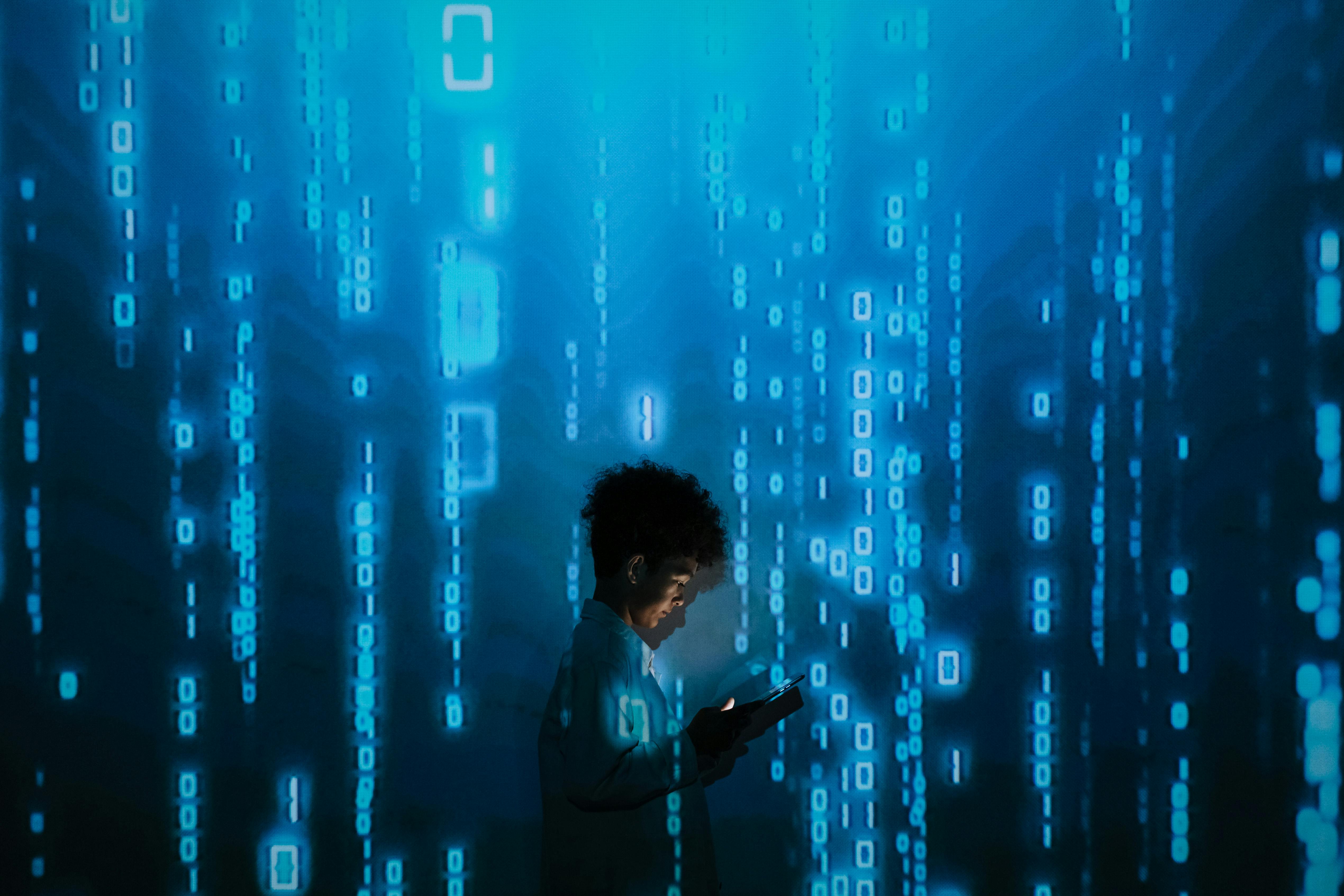 A person with curly hair is silhouetted against a blue digital background featuring cascading binary code (0s and 1s). They are looking down at a mobile device in their hands, which illuminates their face. The visual suggests themes of technology, digital immersion, and the intersection of humanity with data.: Photo by Ron Lach : https://www.pexels.com/photo/person-with-smartphone-standing-in-projection-of-zeros-and-ones-9783812/
