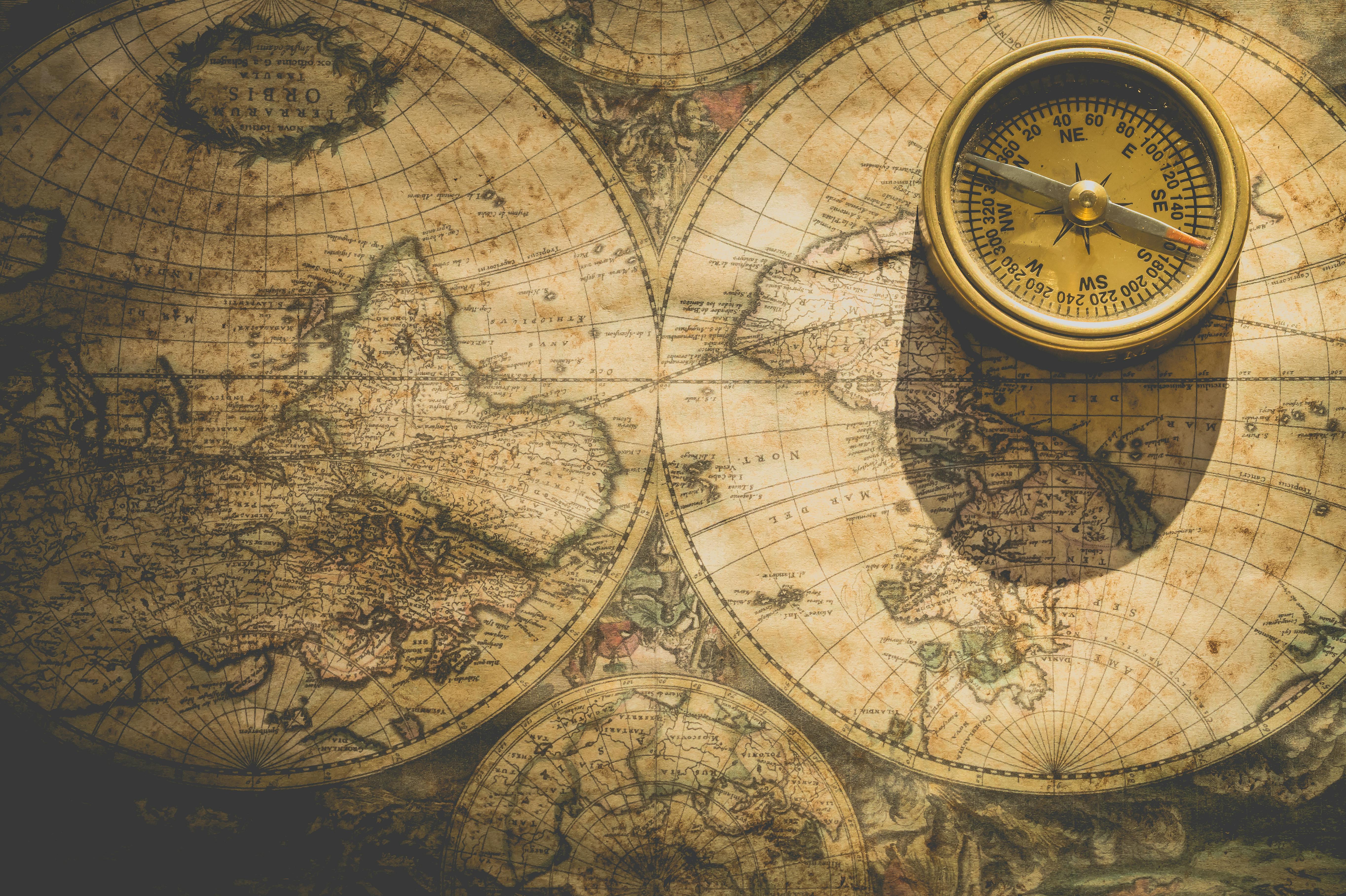 A brass compass with a golden face sits atop an aged, sepia-toned world map. The compass needle points northeast, with detailed degree markings visible around its rim. The underlying map appears to be a vintage double-hemisphere projection showing both the Eastern and Western hemispheres, with ornate cartographic details, grid lines, and geographical features rendered in warm brown and beige tones. The map shows signs of age with weathered edges and faded text, creating an antique, exploratory atmosphere that evokes historical navigation and discovery.